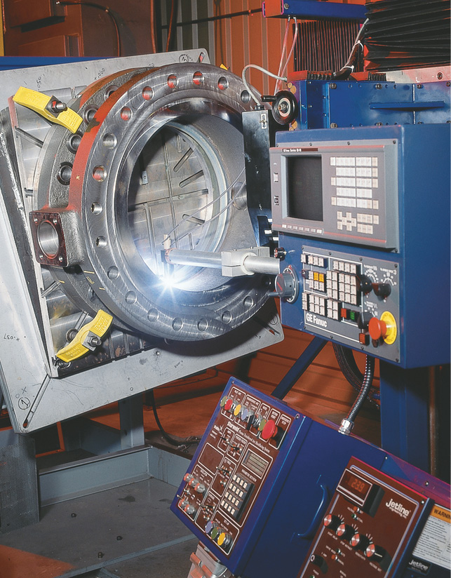 A metalworking machine with a large circular component under processing. Multiple control panels with numerous buttons and dials are visible, and a bright light shines on the component, indicating active machining. The setting appears to be an industrial workshop.