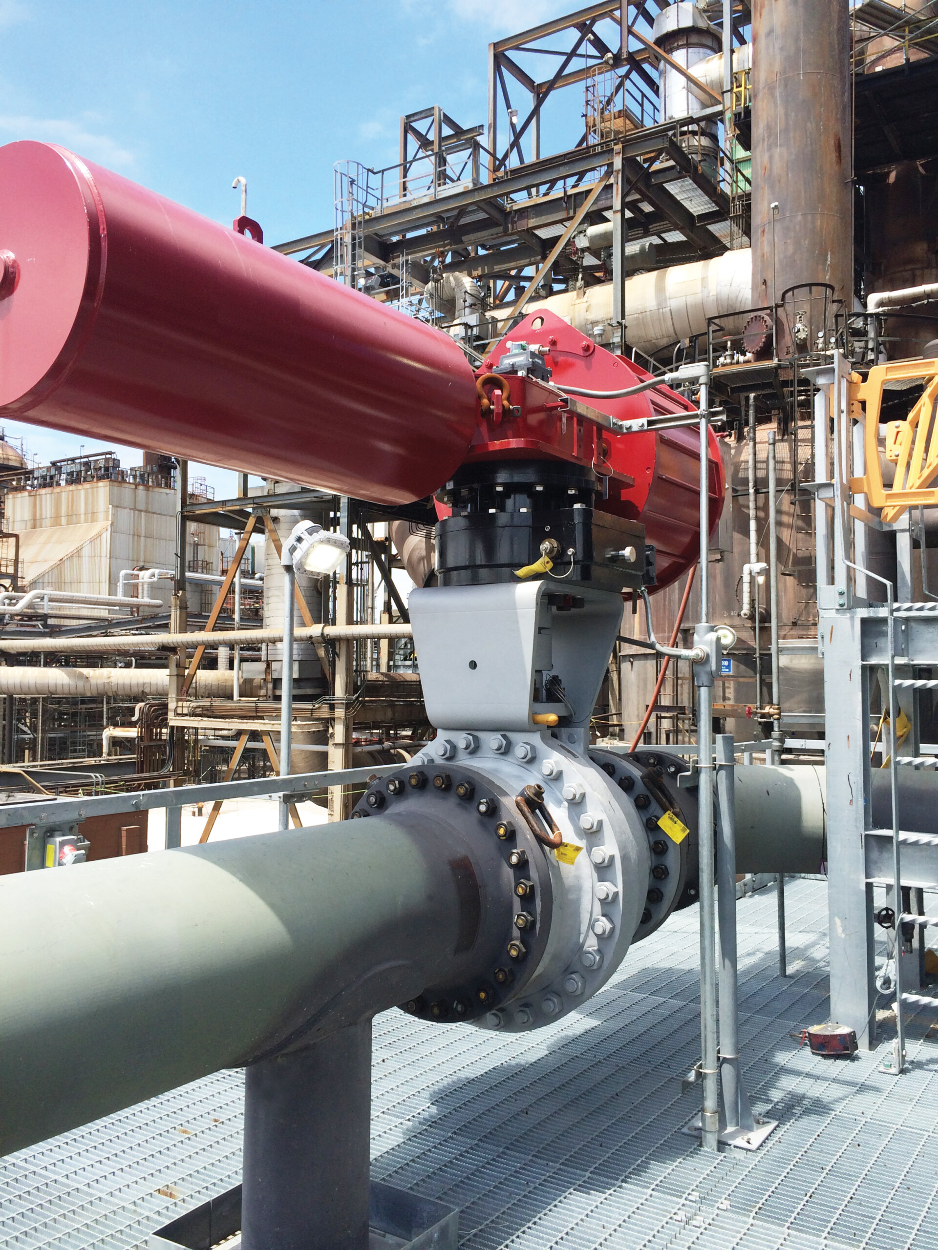 A large industrial valve with a red actuator is installed on a horizontal pipeline at a refinery or processing plant, surrounded by metal structures and pipes under a blue sky.