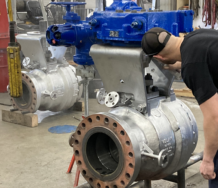 A person in a cap works on a large industrial valve in a workshop. The valve is metallic with large round openings, and a portion is painted blue. Another similar valve is visible in the background. Various tools and machinery are scattered around.