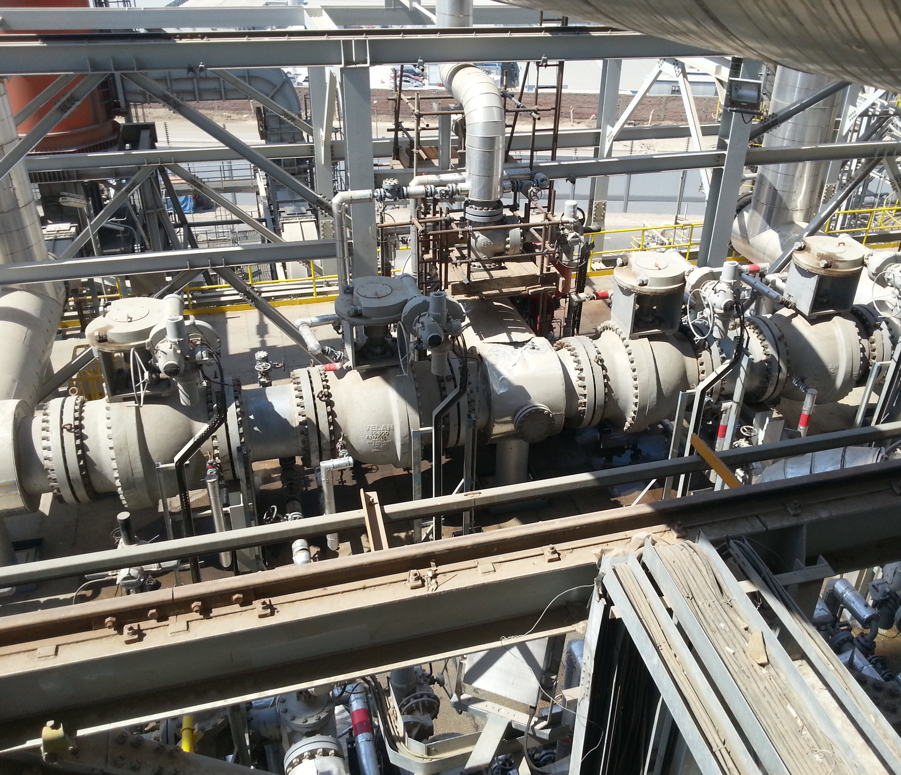 A large industrial facility with interconnected metal pipes and machinery. Valves and gauges are visible, with substantial metal structures and multiple levels. The background shows additional industrial equipment and a glimpse of outdoor surroundings.