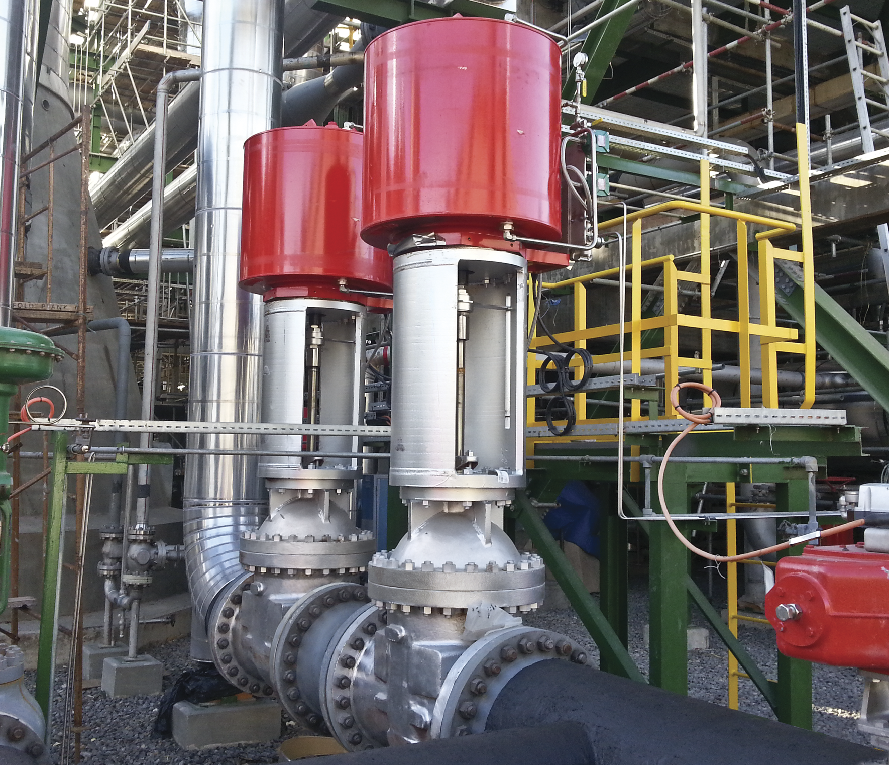 Two large industrial valves with red actuators installed on a pipeline within a complex facility. The setting includes various steel pipes, green support structures, and yellow safety railings, suggesting an industrial or manufacturing environment.